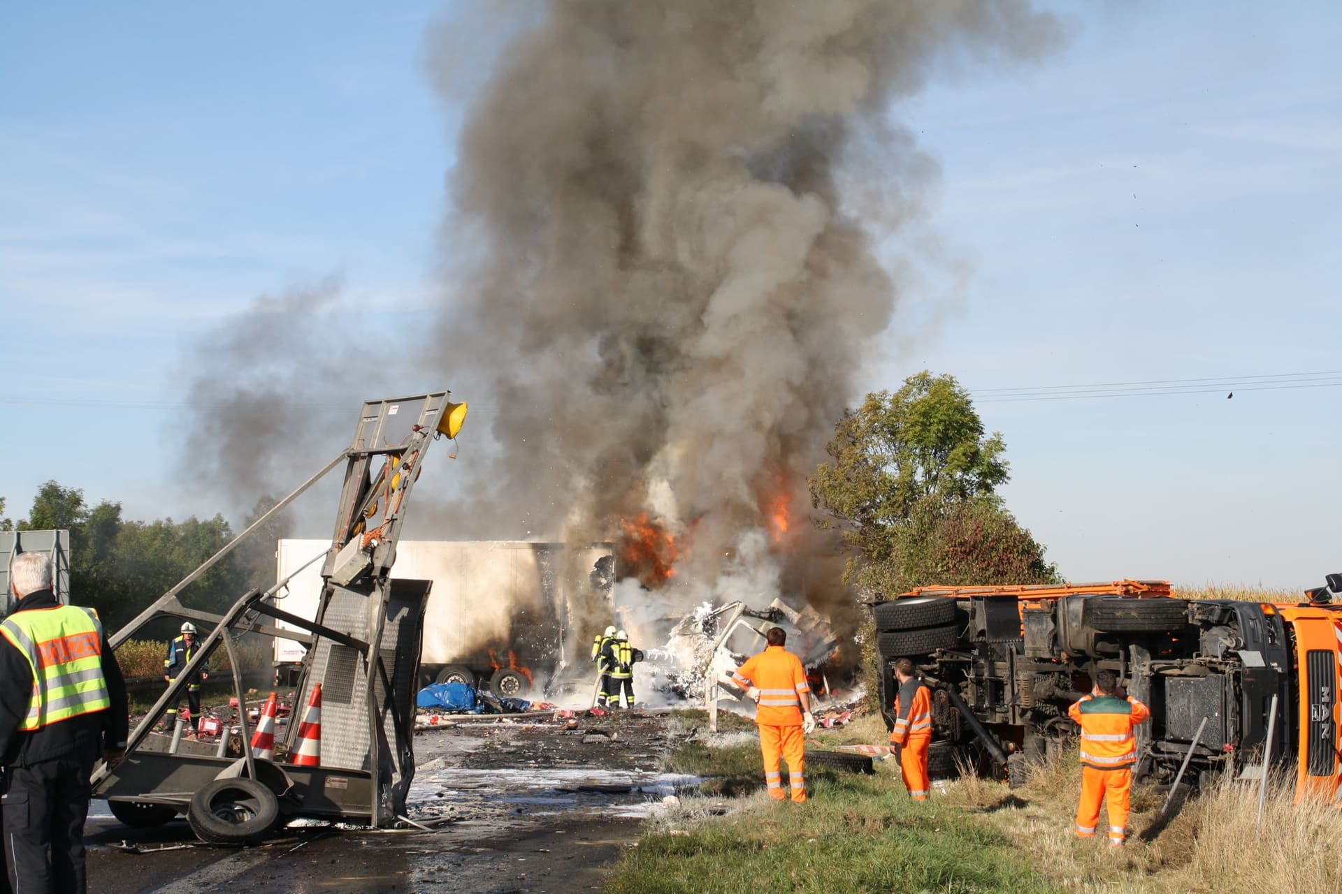 Ein Lastwagen brennt nach einem Auffahrunfall auf der Autobahn, daneben Einsatzkräfte und ein umgestürztes Baustellenfahrzeug